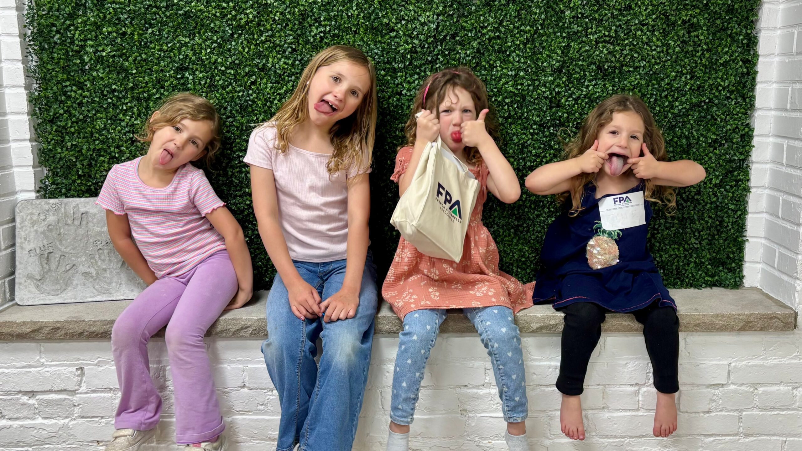 Four kids sit on a stone ledge in front of a green hedge, making goofy faces at the camera.