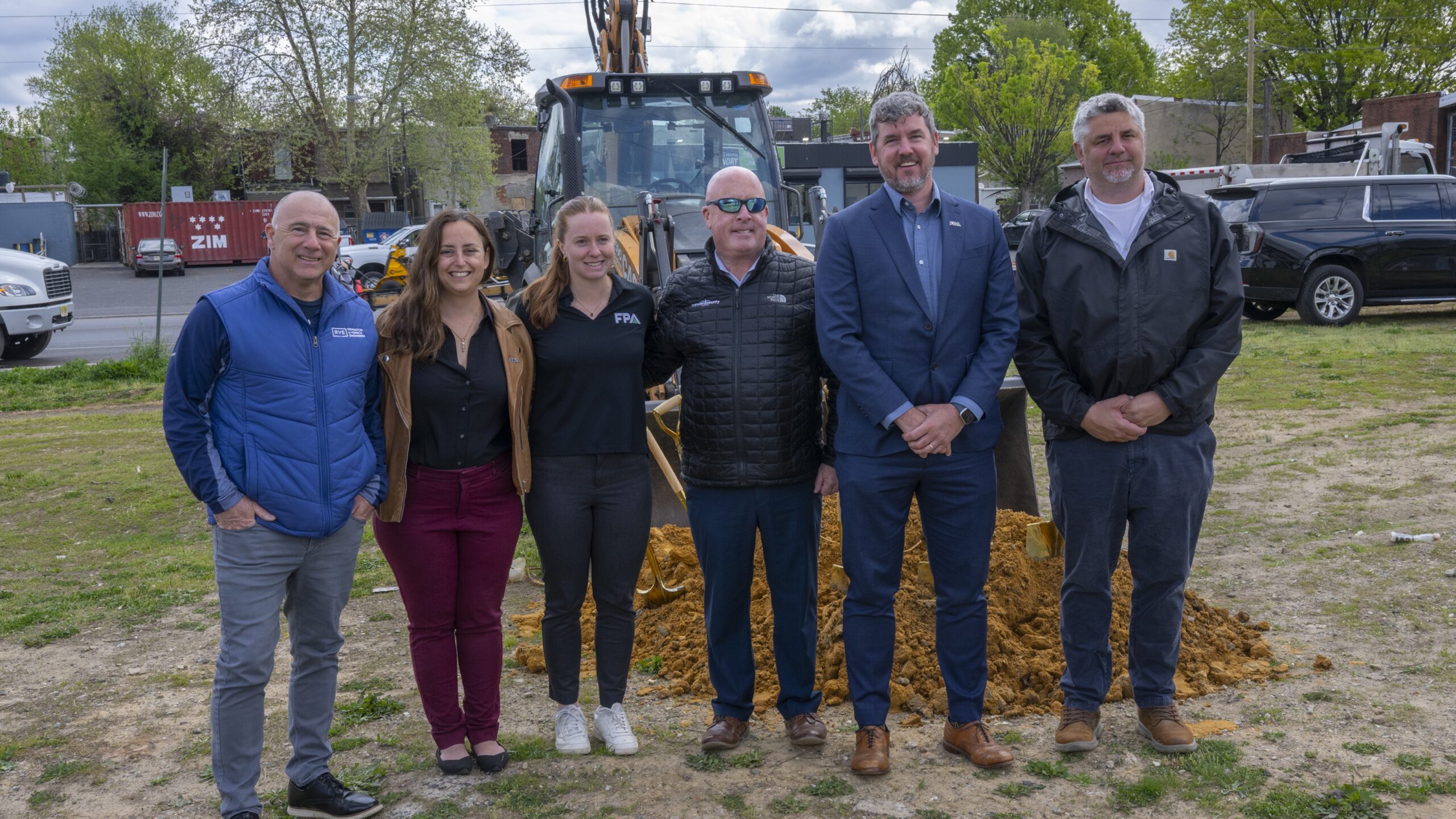 Six adults posing for a group photo at a construction site with a backhoe and dirt pile behind them.