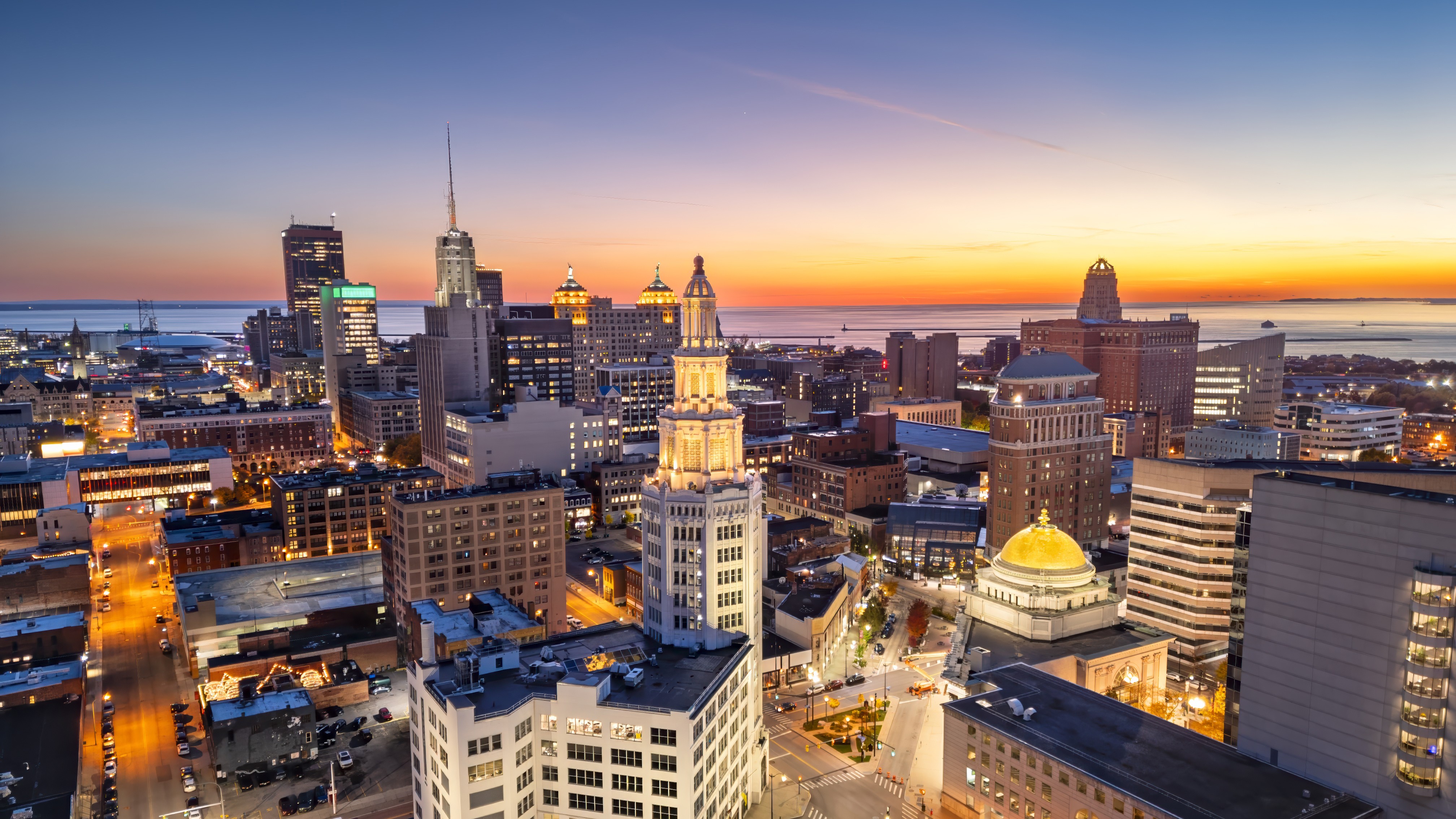 Skyline cityscape at sunset with tall buildings along the waterfront and golden dome in foreground, orange sky fading to blue above sea.