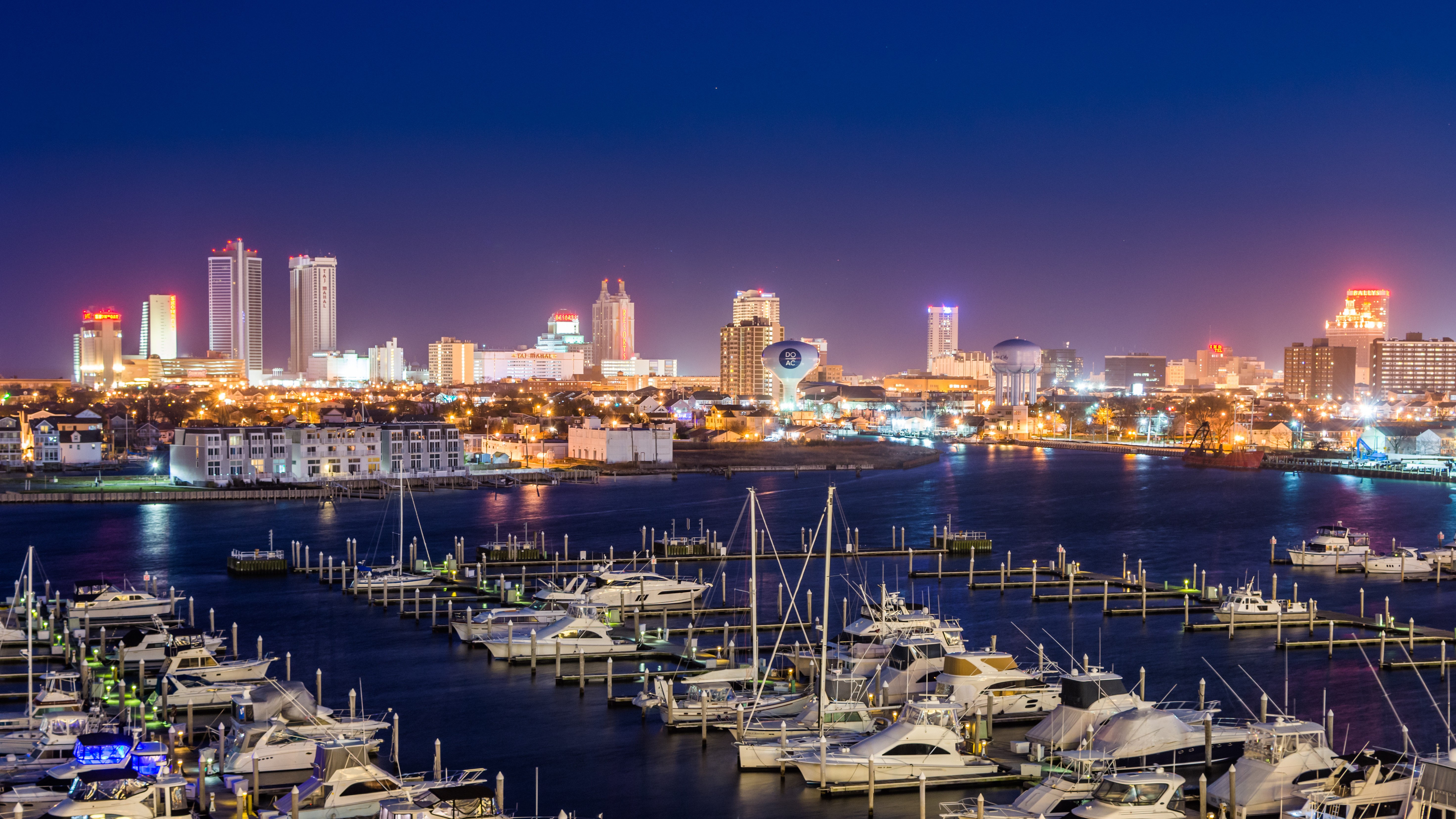 Night view of a waterfront city with illuminated skyscrapers and a marina filled with boats.