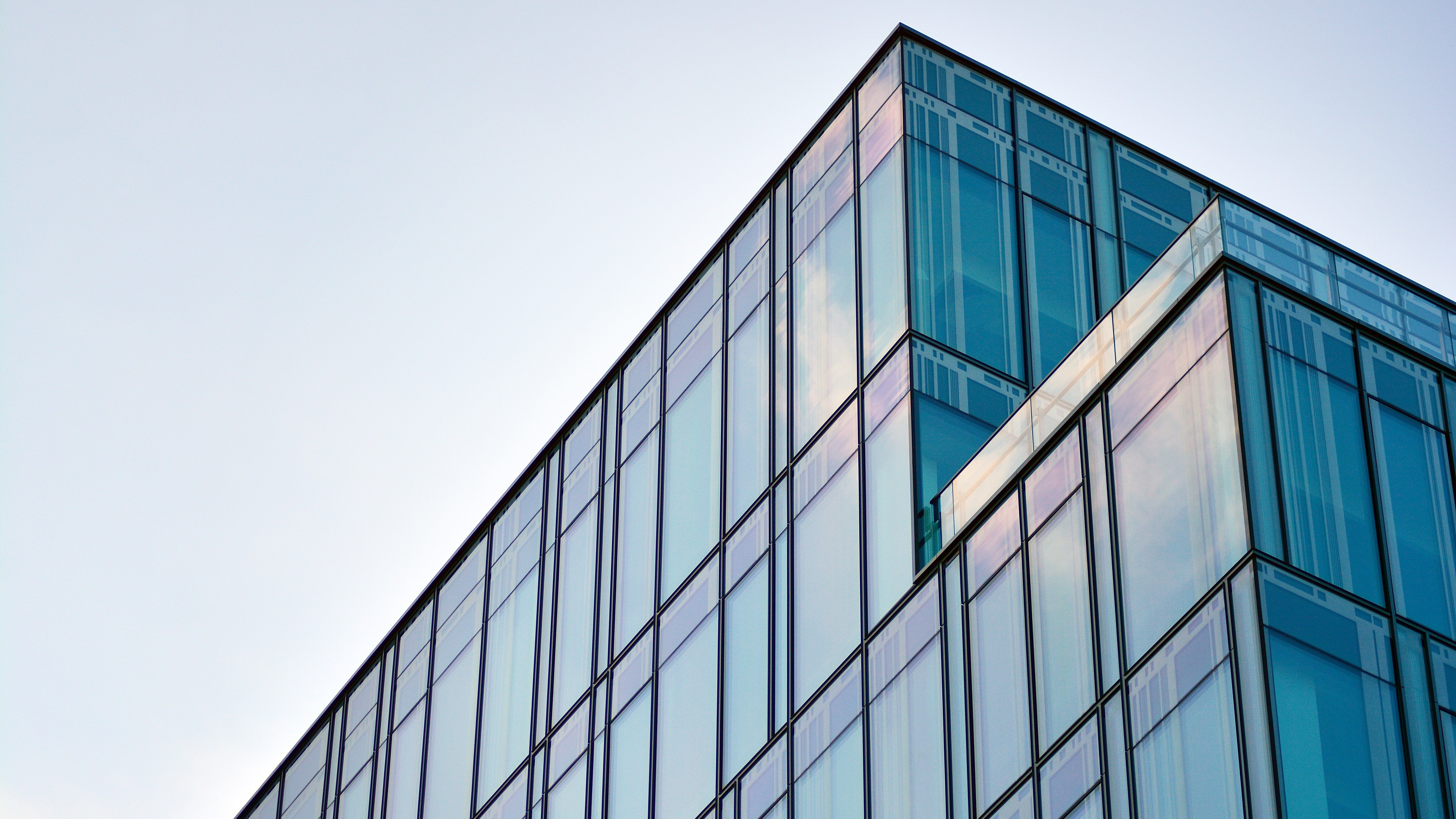 Low-angle view of a glass-office building corner with blue-tinted windows reflecting the sky against a pale blue backdrop.