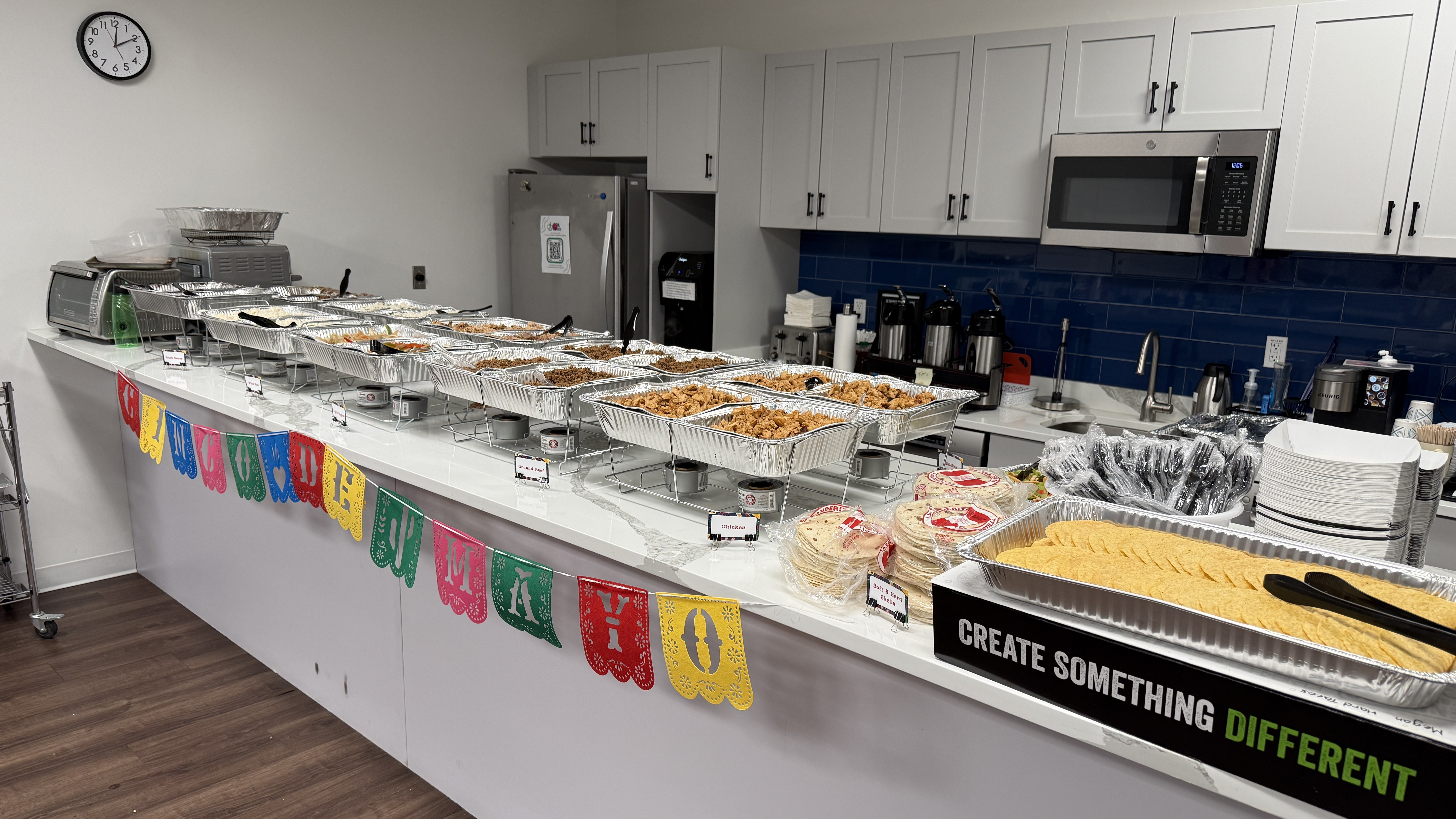 Long buffet counter with aluminum trays of assorted dishes, colorful banner under the counter, in a kitchen or cafeteria setting.