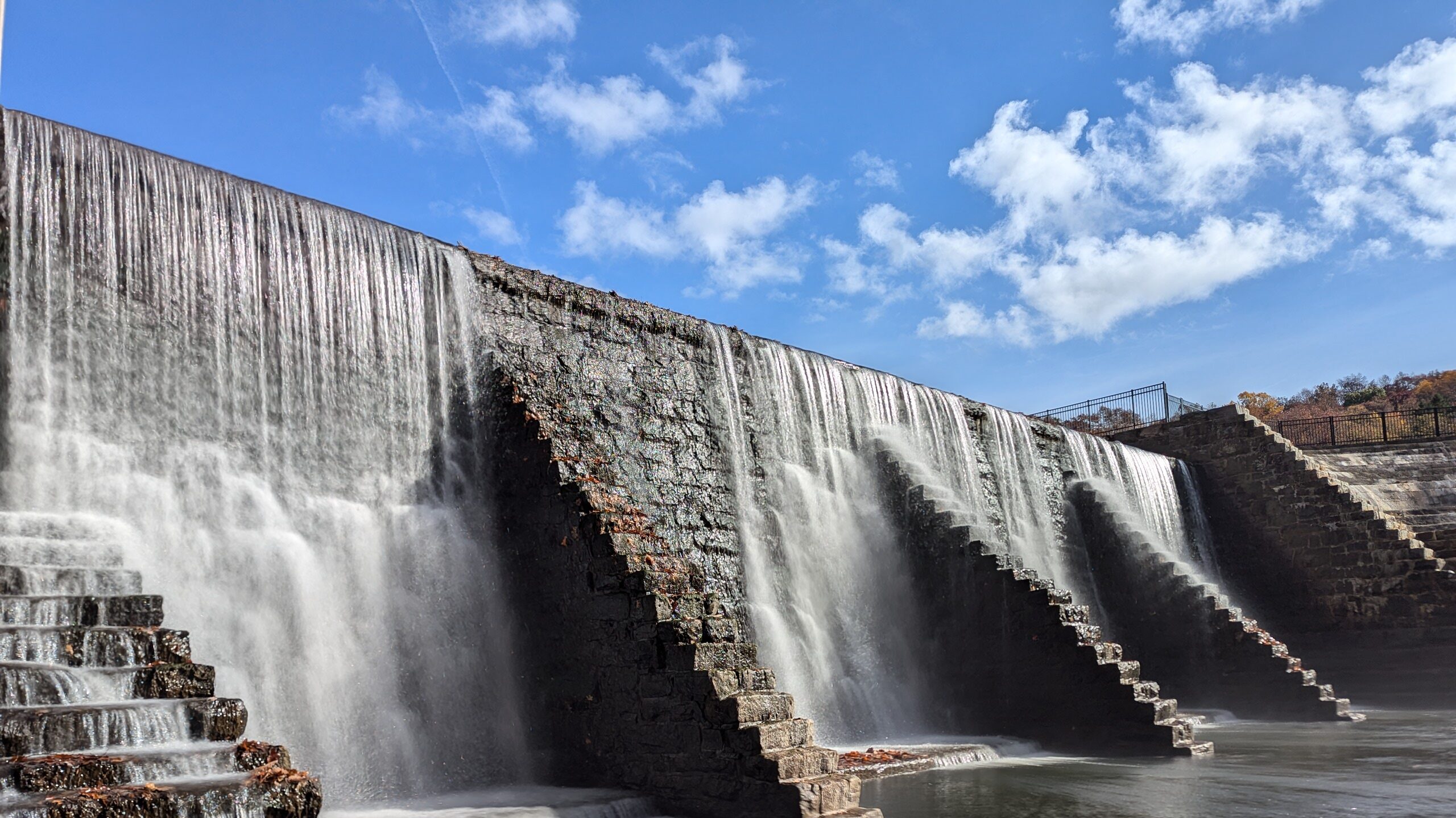 Water cascades over a stepped stone dam on a sunny day, with a blue sky above and trees in the distance.