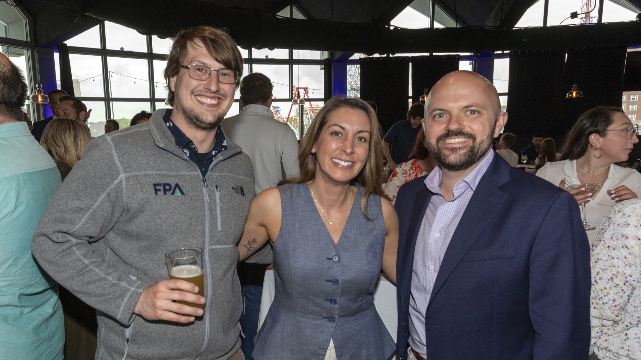 Three friends posing at a crowded indoor party: man left holds a beer, woman center in a blue dress, man right in a navy suit, all smiling.