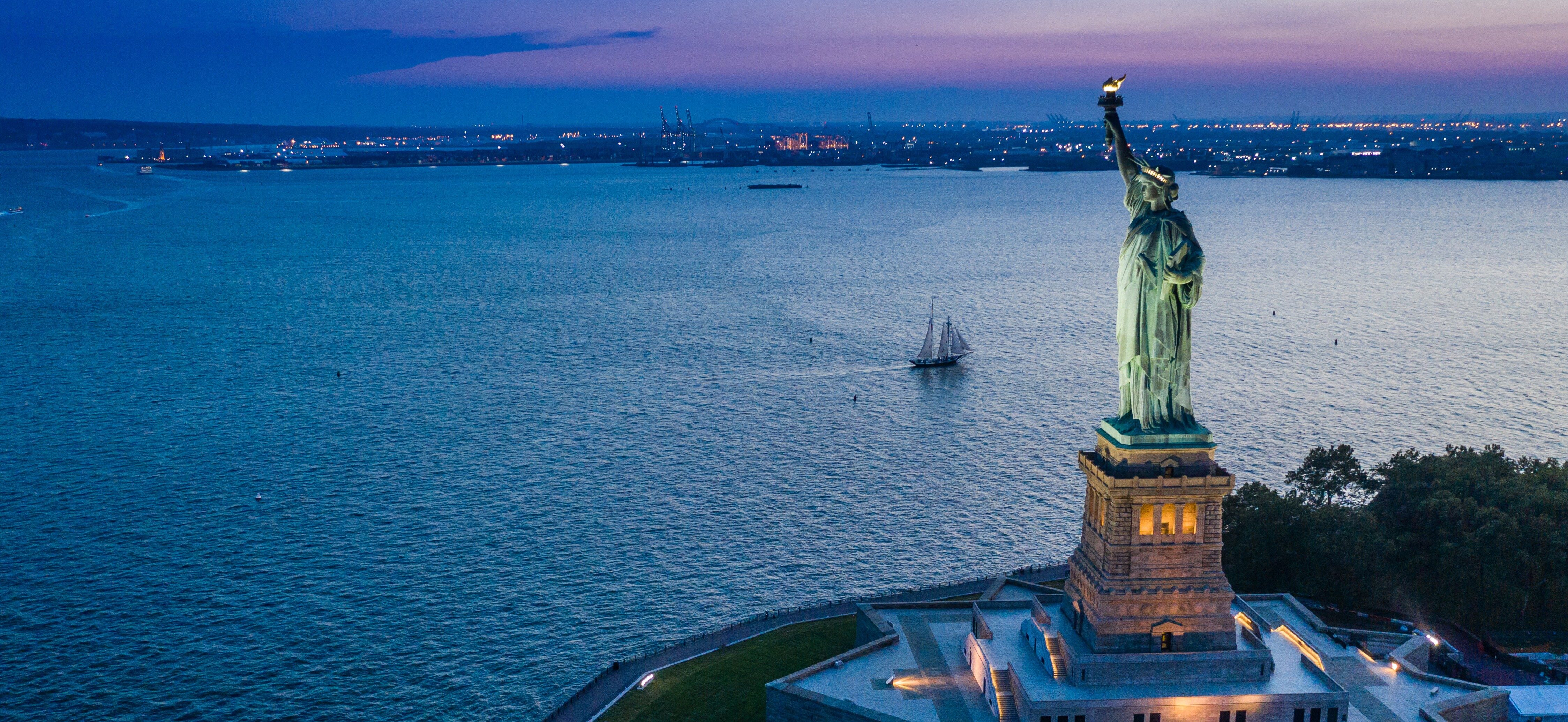 Aerial view of the Statue of Liberty on Liberty Island at dusk, with calm water and distant city lights across the harbor.