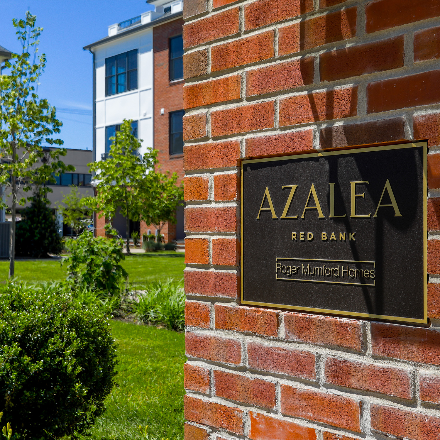 Brick pillar with a gold-framed plaque reading 'AZALEA RED BANK Roger Mumford Homes' and nearby greenery in a sunny development setting
