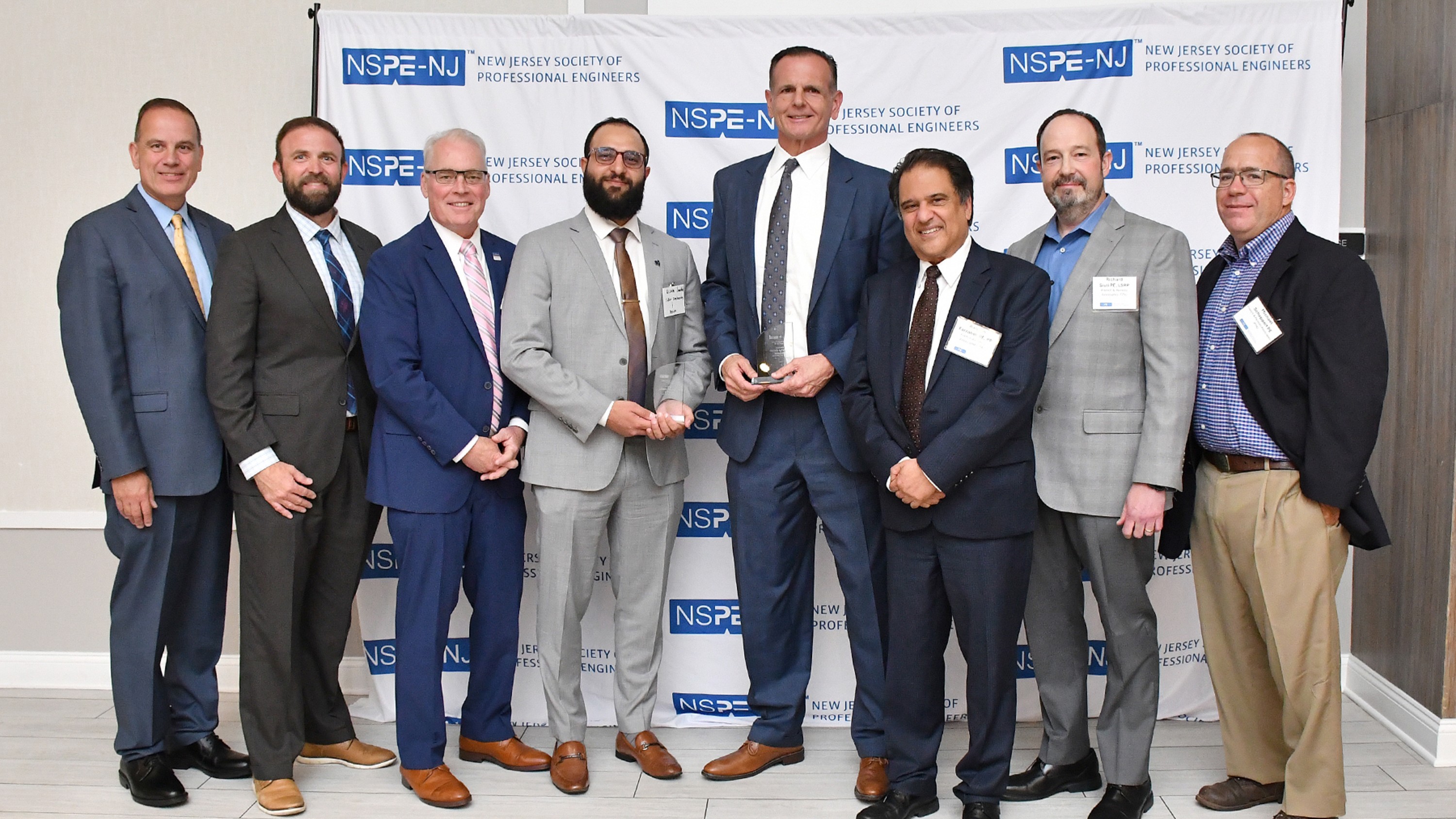 Eight men in business suits pose for a group photo at a New Jersey Society of Professional Engineers event, backdrop displays NSPE-NJ logos.