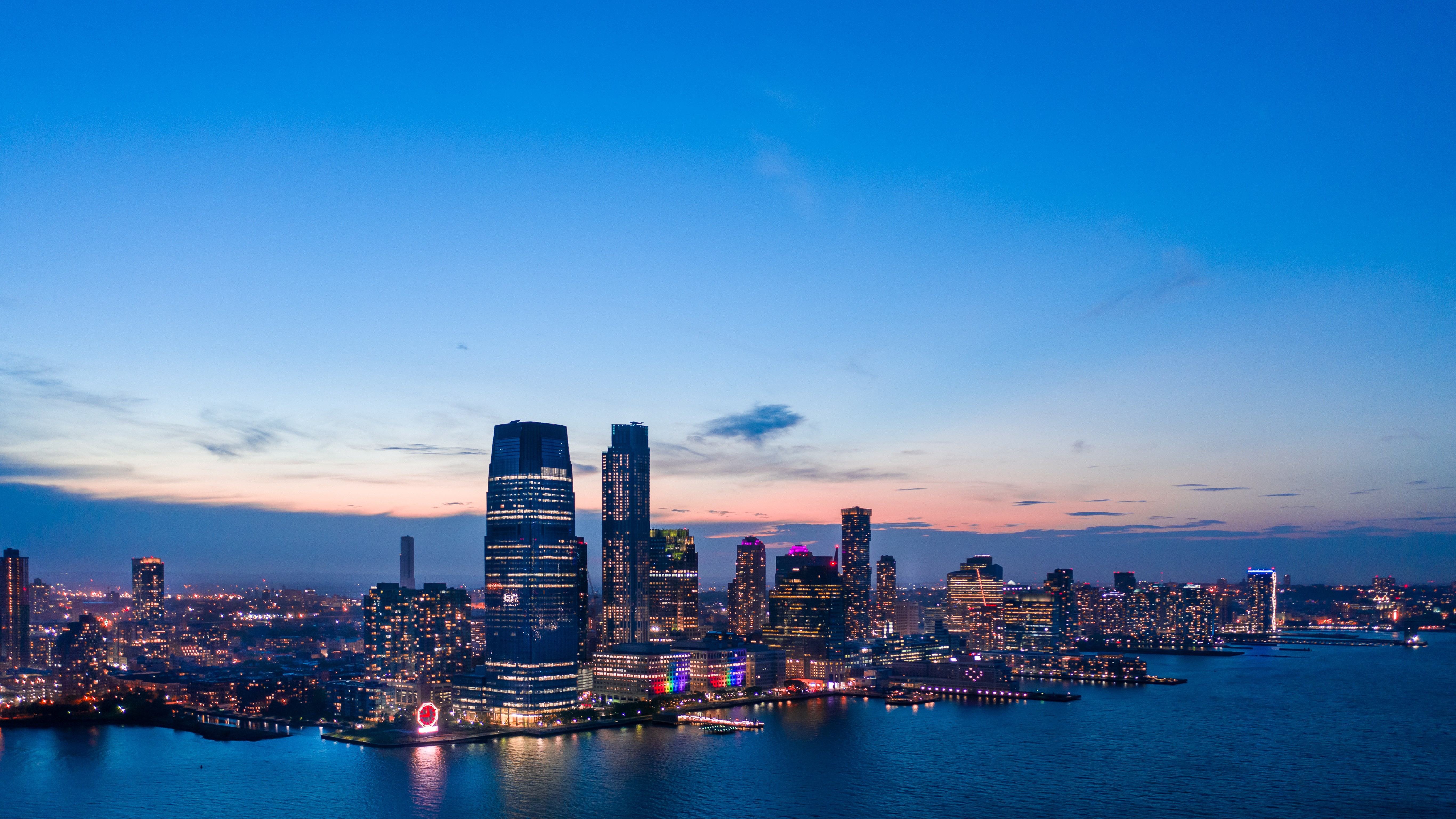 Urban skyline at dusk along a river, with tall glass skyscrapers glowing against a gradient pink-to-blue sky.