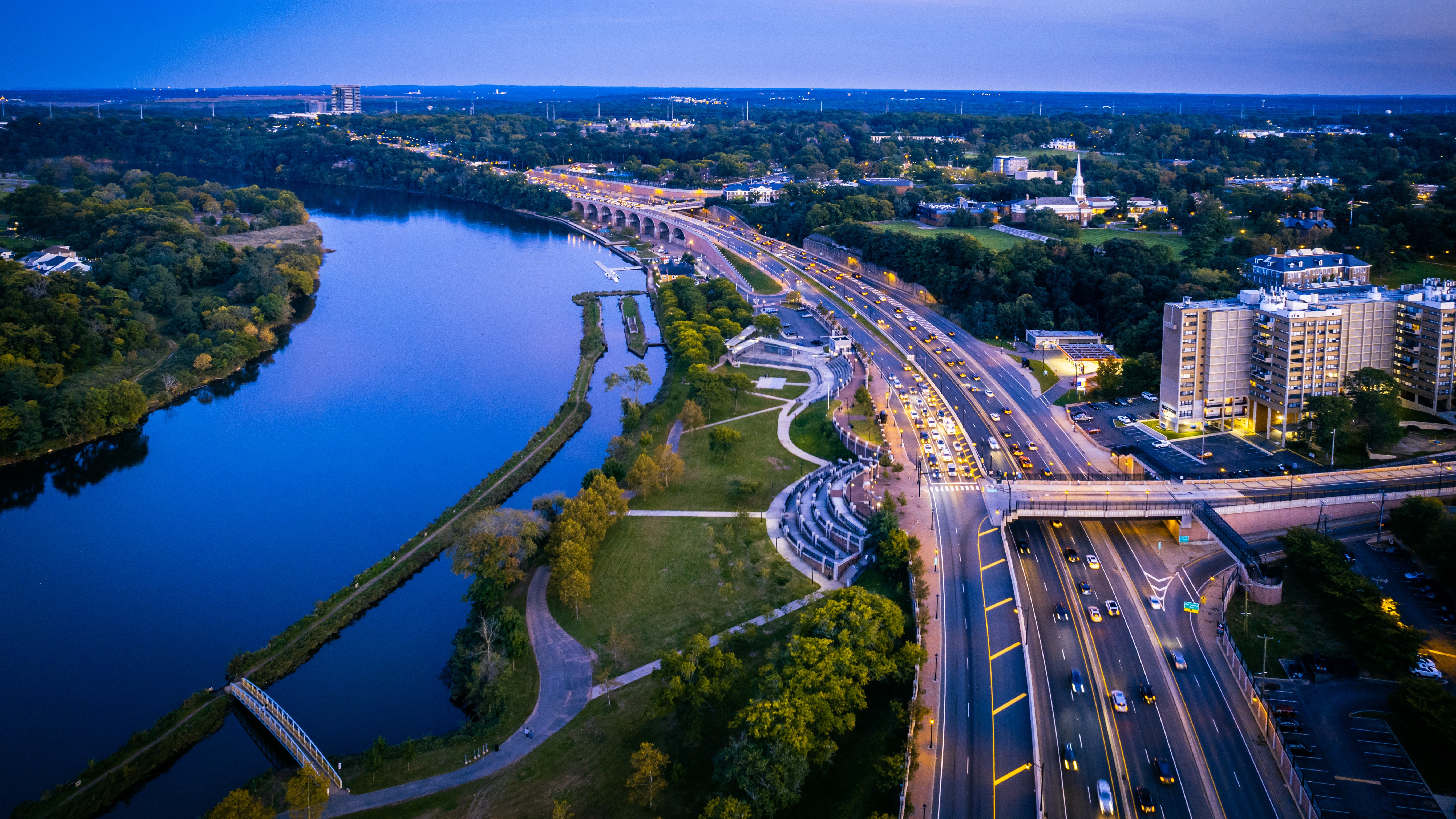 Aerial view of a winding river with a cityscape: highway and bridge along the riverfront, surrounded by trees and buildings on the right.