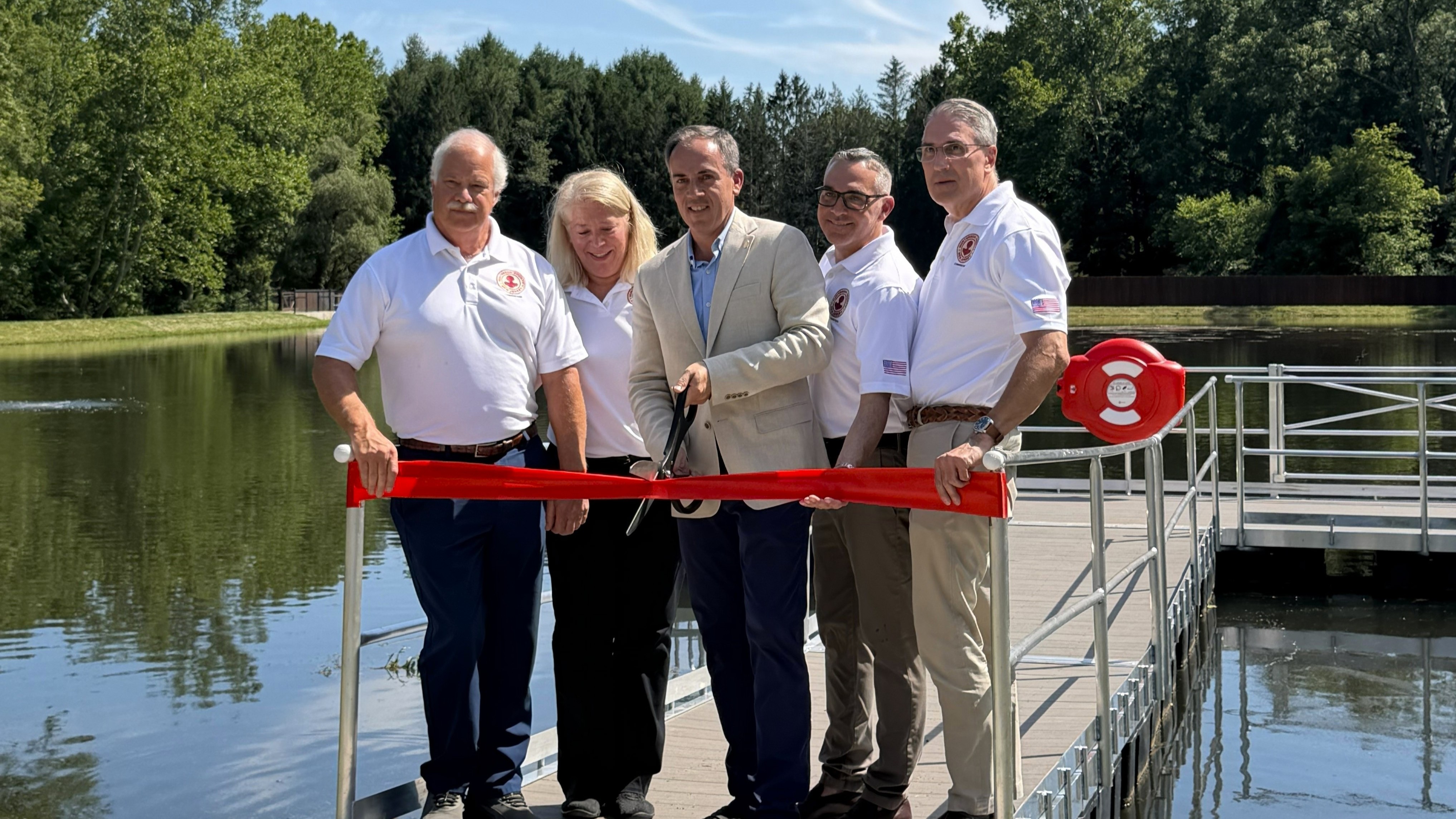 A group of five adults in smart casual clothing cutting a red ribbon on a dock beside calm water.